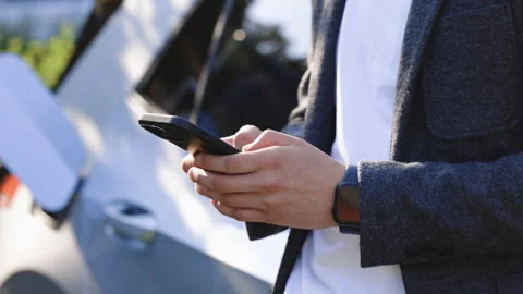 Man holding smartphone while charging car at electric vehicle charging station Stock Footage 249886735