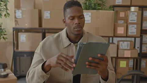 Man holding tablet, hand on forehead, checking shipping labels amid stacked.. Video stock 337434647
