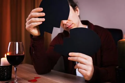 Man holding two halves of a broken black paper heart at a table with wine. .. Stock Photos