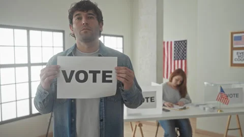 Man holding vote sign in front of a polling table and ballot box inside a b.. Stockbeeldmateriaal 337392473