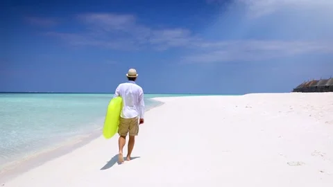 Man holding a yellow floaty walks down a tropical paradise beach Stock Footage 264157304