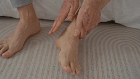 Man holds ankle on bed in studio, hands gripping foot and lower leg with vi.. Stockbeeldmateriaal 337445416