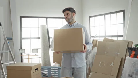 Man holds cardboard box with both hands inside building amid stacked moving.. Stock Footage 339175874