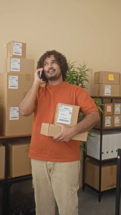 Man holds phone to ear while holding labeled cardboard package amid stacked.. Stockbeeldmateriaal 337395407