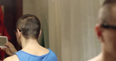 A man holds a small mirror in his hands and shaves the hair on his head. Stock Footage 103805963
