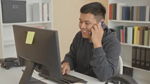 Man holds smartphone to ear while typing on computer at building desk, smil.. Stock Footage 347803441