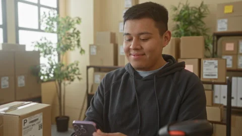 Man holds smartphone in hands and smiles over shipping parcels inside a bui.. Stockbeeldmateriaal 337353774