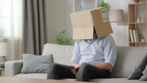 Man holds smartphone while sit on sofa with box on his head, using parcel Stock Footage 169017466