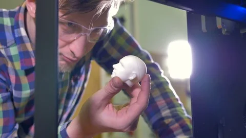 A man holds a tiny doll head sample and smiles at a working 3d-printer. 4K. Stock Footage 78352770