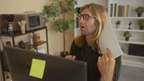 Man holds white document to forehead at desk in cluttered office with compu.. Stock Footage 313462415