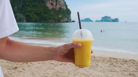 Man holds yellow mango smoothie in plastic cup with a straw on tropical sandy Stock Footage 282919880