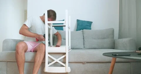 A man in home clothes assembles a chair with a screwdriver by himself Stock Footage 160487968