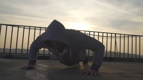 Man with hood over head doing push ups on terrace, sun shining in the background Stock Footage 98485109