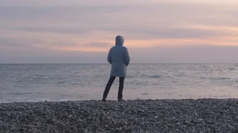 A man in a hooded jacket in cold weather stands on the beach and looks at the 스톡 동영상 120843862