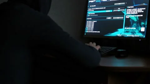 A man in a hooded sweatshirt is sitting at a computer in the dark. light from Video stock 165833235