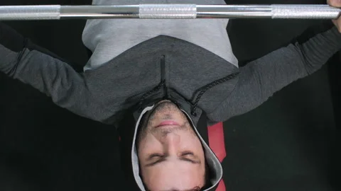 Man in hoodie is doing bench press exercise lifting barbell in gym, top view. Video stock 231336883