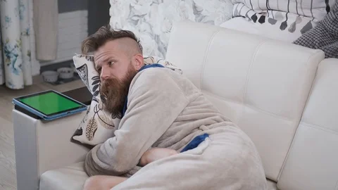 Man in housecoat is lying on sofa next to chromakey tablet Stock Footage 127886044