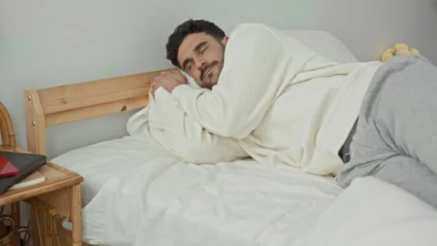Man hugging pillow and lying on his side on a bed, eyes closed, wearing swe.. Stock Footage 339176371