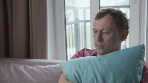 A man immersed in his thoughts sits on a sofa clutching a pillow to himself Stock Footage 154433777