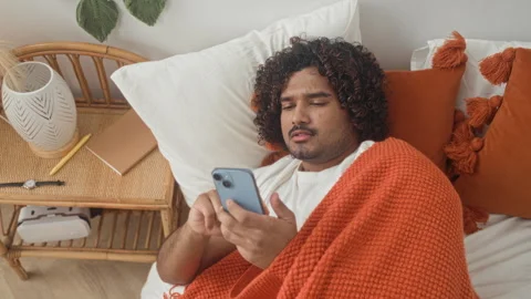 Man indian young lying in bed under an orange blanket holding smartphone an.. Stock Footage 337401567