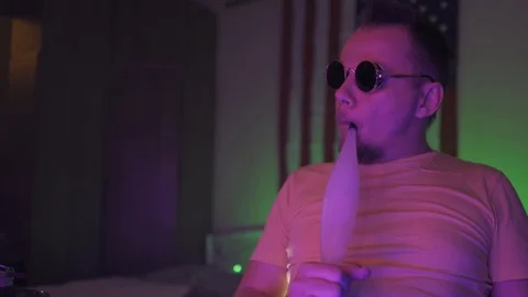 Man Inhaling from a Hookah dancing sitting on a chair in neon light room wearing Video stock 130064311