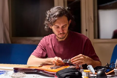 Man inspecting small guitar electronics component at cluttered workbench in dim Fotos de archivo