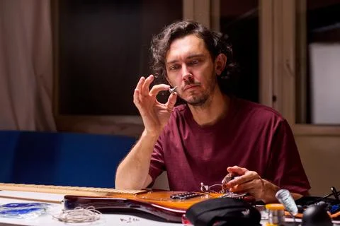 Man inspecting small guitar electronics component at cluttered workbench in dim Fotos de archivo