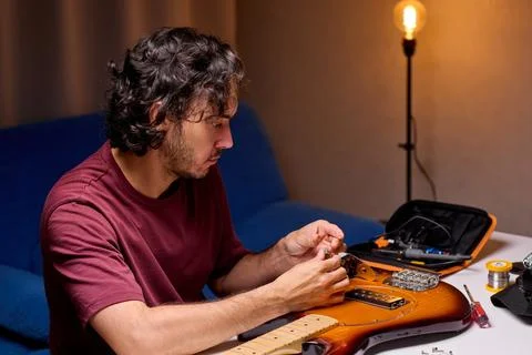 Man inspecting small guitar electronics component at cluttered workbench in dim Fotos de archivo