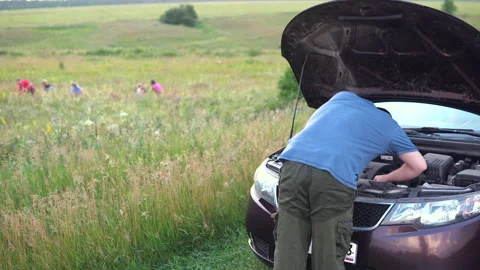 A man inspects a car engine while a family walks in a field. Car breakdown Stock Footage 134492874