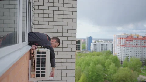 A man installs an external air conditioning unit outside the window of an Stock Footage 195936228