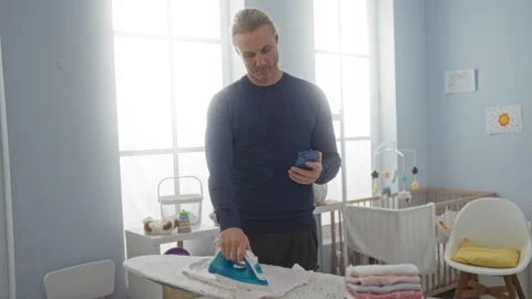 Man ironing clothes while checking phone in a bright laundry room with iron.. Stock Footage 310641237