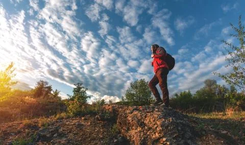 A man in a jacket and with a backpack goes over a large stone at sunset Stock Photos