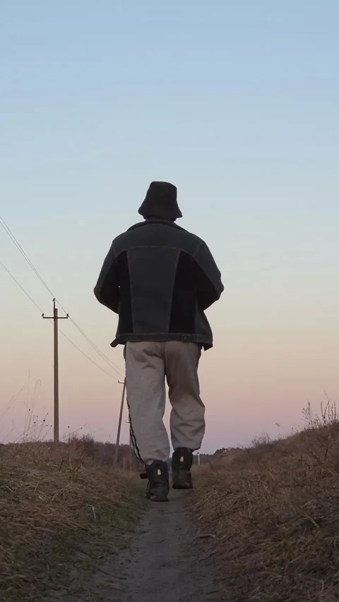 Man in jacket and grey sweatpants walking away along narrow path against ev.. Vídeo Stock 333791322
