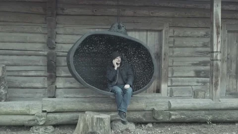 Man in jacket and hat speaks on phone sitting on large swing Stock Footage 147171367