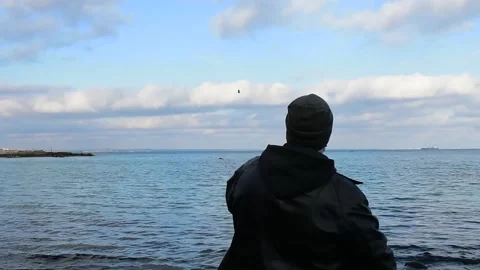 A man in a jacket and a hat throws stones into the water from the shore Stock Footage 141056619