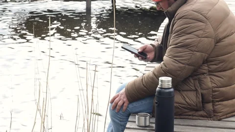A man in a jacket and orange hat sits on a wooden jetty   Stock Footage 275132477