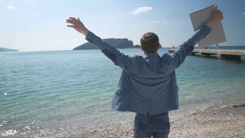 A man in a jacket with a laptop stands on the beach with his arms outstretched 스톡 동영상 151514164