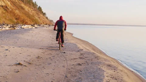 Man in jacket rides bicycle along sandy river bank Video stock 231482099