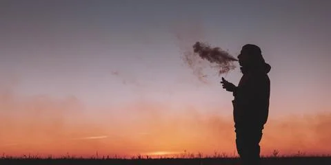 A man in a jacket smokes an electronic cigarette outdoors. Vaping on a sunset Stock Photos