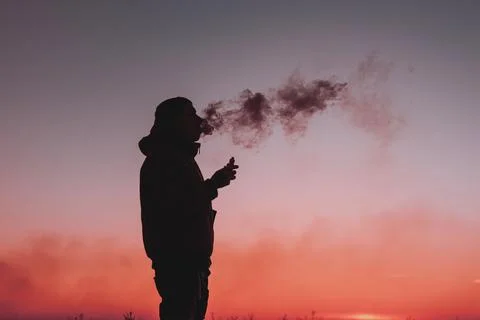 A man in a jacket smokes an electronic cigarette outdoors. Vaping on a sunset Stock Photos