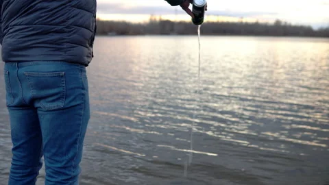A man in a jacket stands on the banks of a river, lake and pours a drink into Stock Footage 142895280