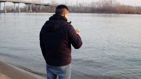 A man in a jacket stands on the riverbank, drinks a drink and throws a can into Video stock 142593463