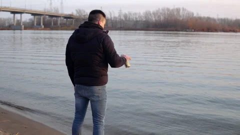 A man in a jacket stands on the riverbank, drinks a drink and throws a can into Stock Footage 142593640