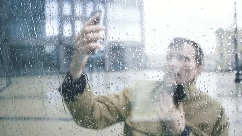 Man in jacket using smartphone under rain. View through window with raindrops Stock Footage 79414873