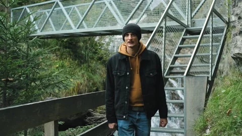 Man in jeans jacket walking on bridge over wild mountain waterfall Stock Footage 256333495