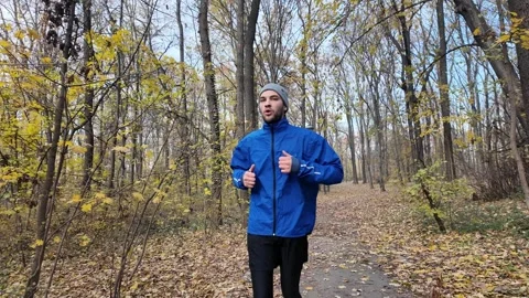 Man jogging in autumn forest wearing blue jacket and beanie, training on a leaf Stock Footage 316845663