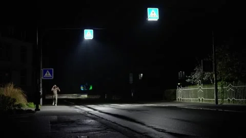 A man is jogging down the street at night, along a dark street illuminated by Stock Footage 308928386