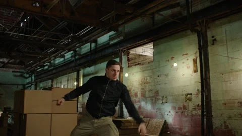 Man jumps over stack of boxes and is chased by another man in dirty warehouse wi Stock Footage 199455365