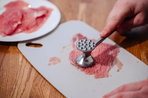 A man in the kitchen is beating red meat. A piece of meat is on a cutting board. Stock Photos
