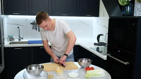 Man in Kitchen Preparing Dinner. Cuts Chicken Breast with Knife. Cooking Salad Stock Footage 147237415